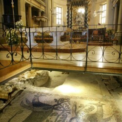 Interno della parte settecentesca dell’Abbazia di Fruttuaria con l’area presbiteriale superiore rialzata e sullo sfondo l’altare a colonne tortili con il gruppo scultoreo dell’Assunta; al livello inferiore il mosaico pavimentale dell’antica Abbazia benedettina, fine XI-inizio XII secolo, raffigurante due maestosi grifoni rampanti separati da un tralcio vegetale, con cornice a tessere nere e inserti rossi di terracotta.