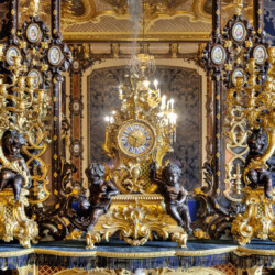 La parure da camino composta da un orologio e due candelabri, che decora la Sala di Trattenimento della Regina, o Salotto Blu, nell’Appartamento di Vittorio Emanuele II, fu acquistata dalla Real Casa presso il mercante Tommaso Haid di Torino. Realizzato intorno al 1850 a Parigi, l’orologio, decorato con putti che alludono alla poesia e alla geometria, è firmato dall’orologiaio Paul Garnier e, insieme ai candelabri, si inserisce perfettamente nell’arredo della sala in stile Napoleone III, o Secondo Impero.