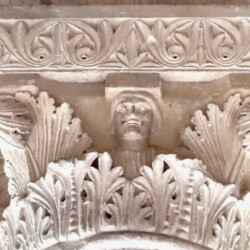 L’apparato decorativo delle chiese romaniche è ricco di immagini simboliche, spesso riconducibili a tradizioni precristiane, ma anche di figure inserite in contesti inconsueti. Non poche sono le ‘insolite presenze’ che si possono scorgere all’Abbazia di Vezzolano, dove, ad esempio, in uno dei capitelli del chiostro, opera risalente al 1180 circa, tra le foglie di acanto si affaccia un busto maschile che indossa una tunica a fitte pieghe e sfoggia un’acconciatura con capelli a caschetto.